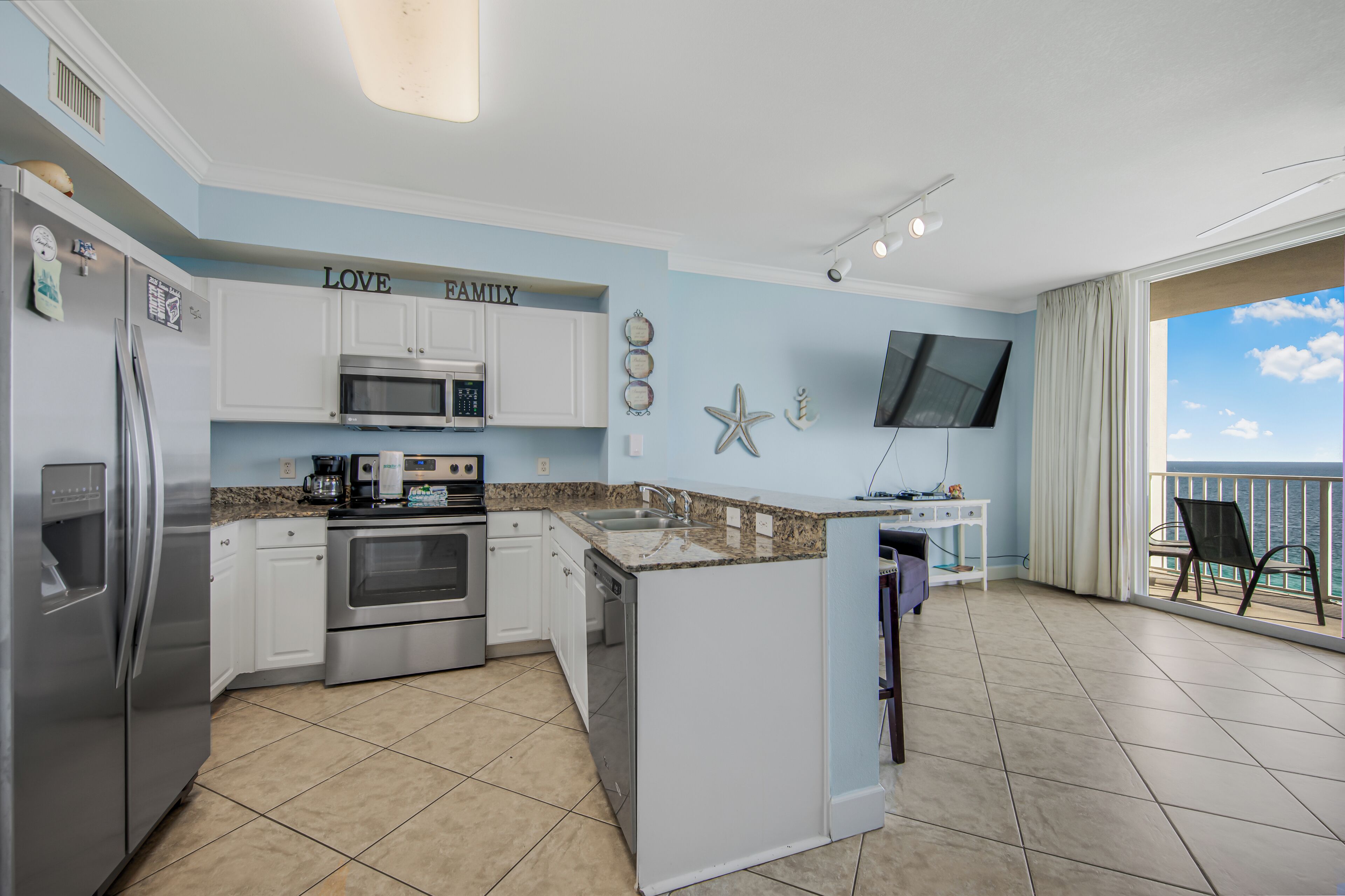 A kitchen with white cabinets, a microwave, an oven, and a coffee maker. There is a small island with a sink and a television mounted on the wall.