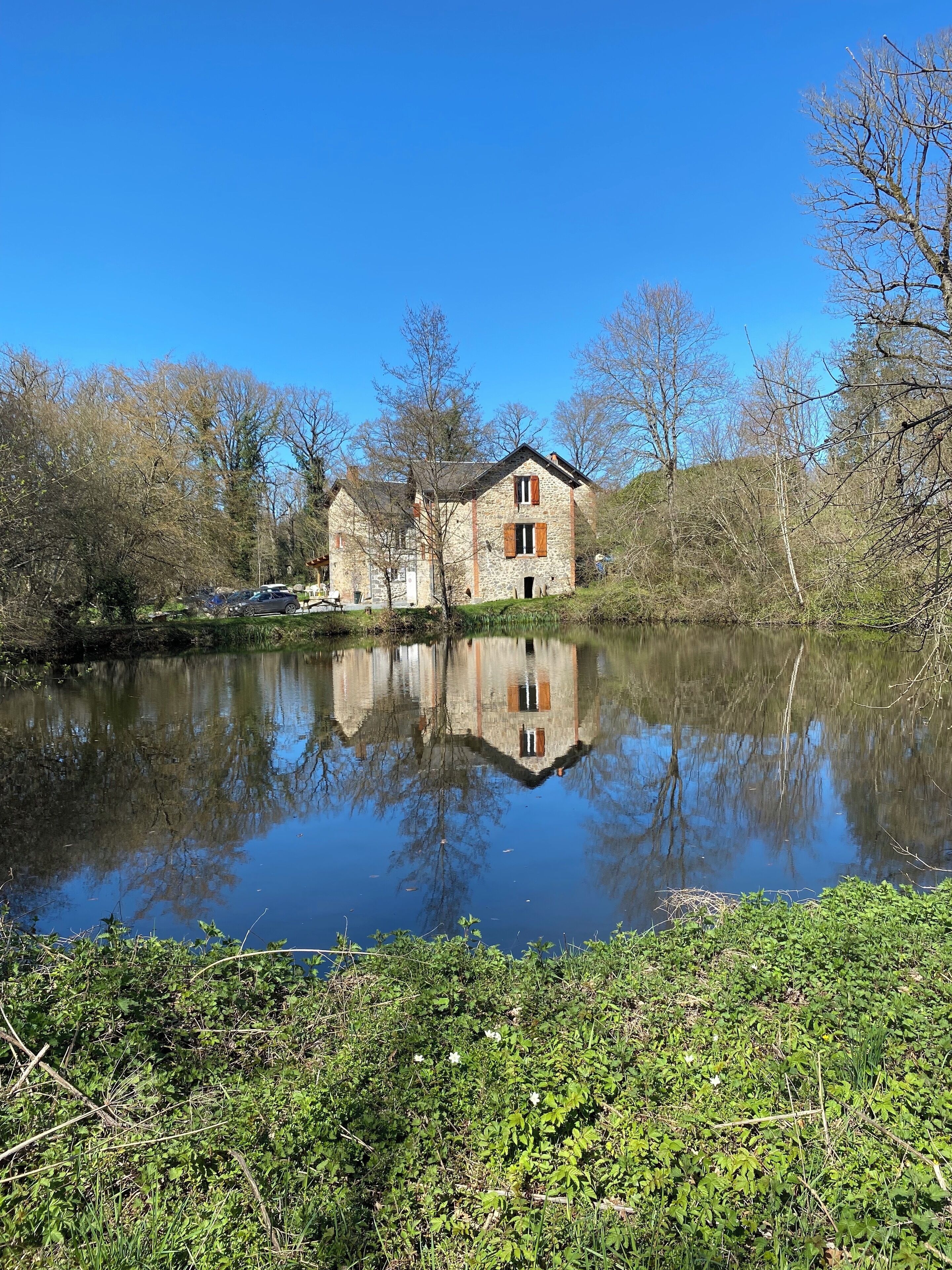 Un bâtiment en pierre qui se reflète dans l’eau, entouré d’arbres et de végétation.