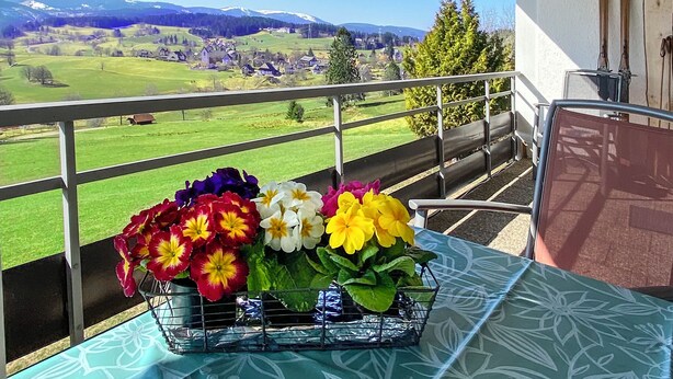 A balcony with a table holding a flower arrangement, a chair, and a view of a green landscape with houses and mountains.