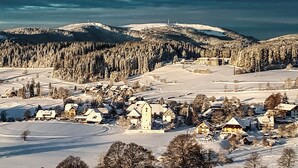A snow-covered village with a church and residential buildings, surrounded by forested hills.