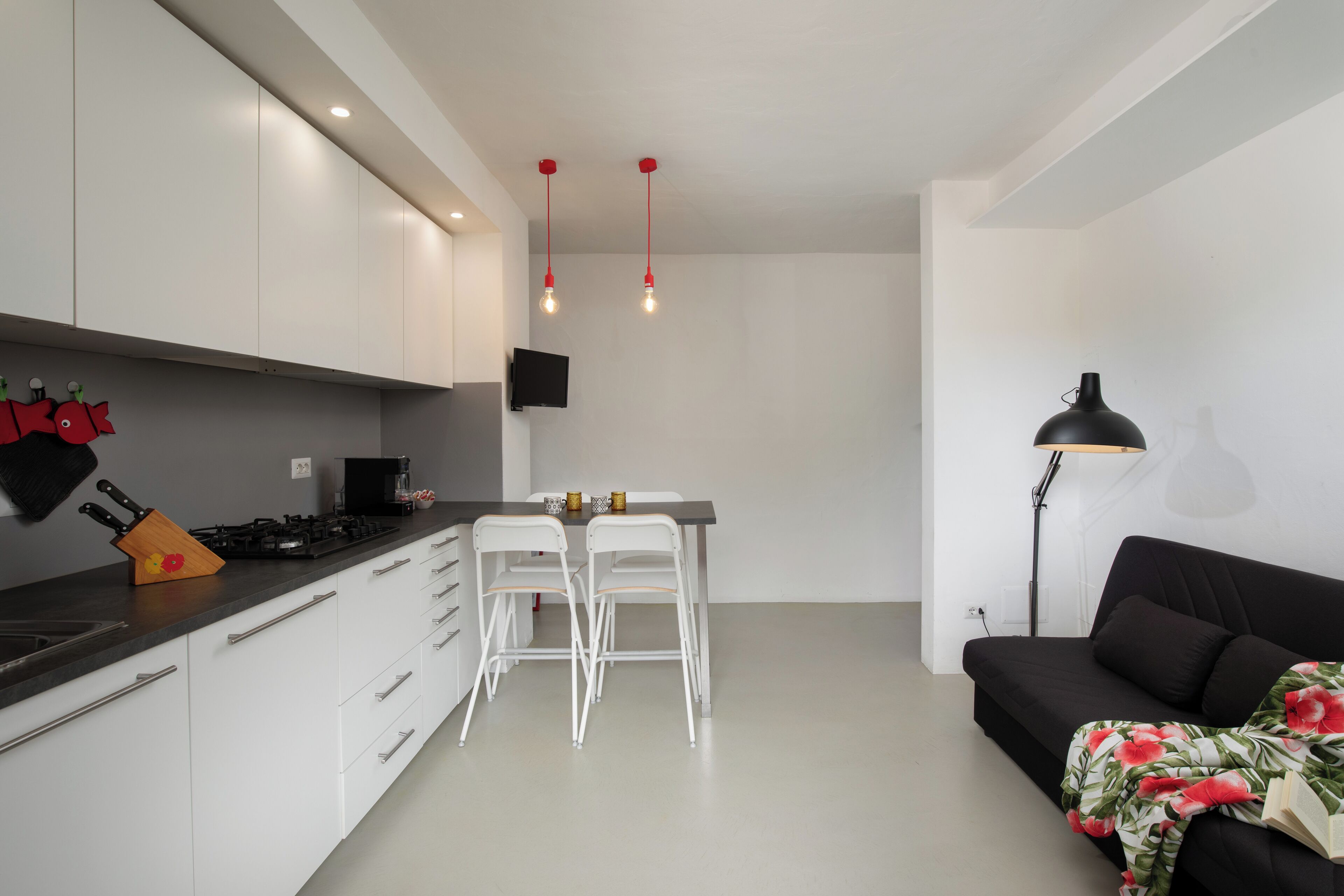 Eine moderne Küche mit weißen Schränken, einer schwarzen Arbeitsplatte und einem Essbereich mit zwei weißen Stühlen. Es gibt ein schwarzes Sofa mit einem floralen Überwurf und eine Stehlampe.