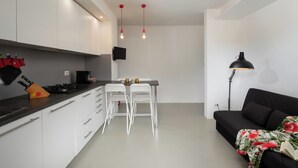 Eine moderne Küche mit weißen Schränken, einer schwarzen Arbeitsplatte und einem Essbereich mit zwei weißen Stühlen. Es gibt ein schwarzes Sofa mit einem floralen Überwurf und eine Stehlampe.