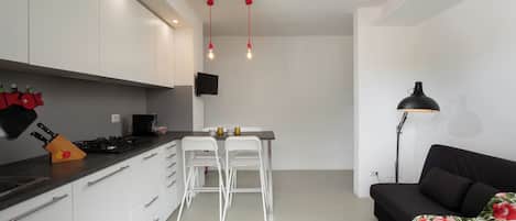 A modern kitchen with white cabinets, a black countertop, and a dining area with two white chairs. There is a black sofa with a floral throw blanket and a floor lamp.