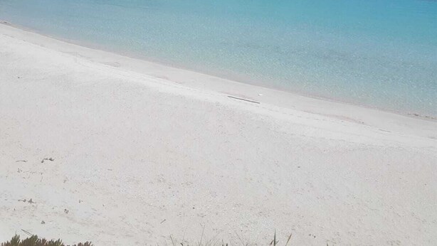 Ein Strand mit klarem türkisblauem Wasser, weißem Sand und im Hintergrund Felsformationen.