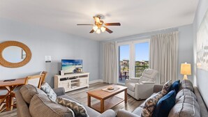 Ein Wohnzimmer mit Deckenventilator, Fernseher, einer Sofagarnitur, einem Couchtisch und Blick ins Freie durch ein großes Fenster.