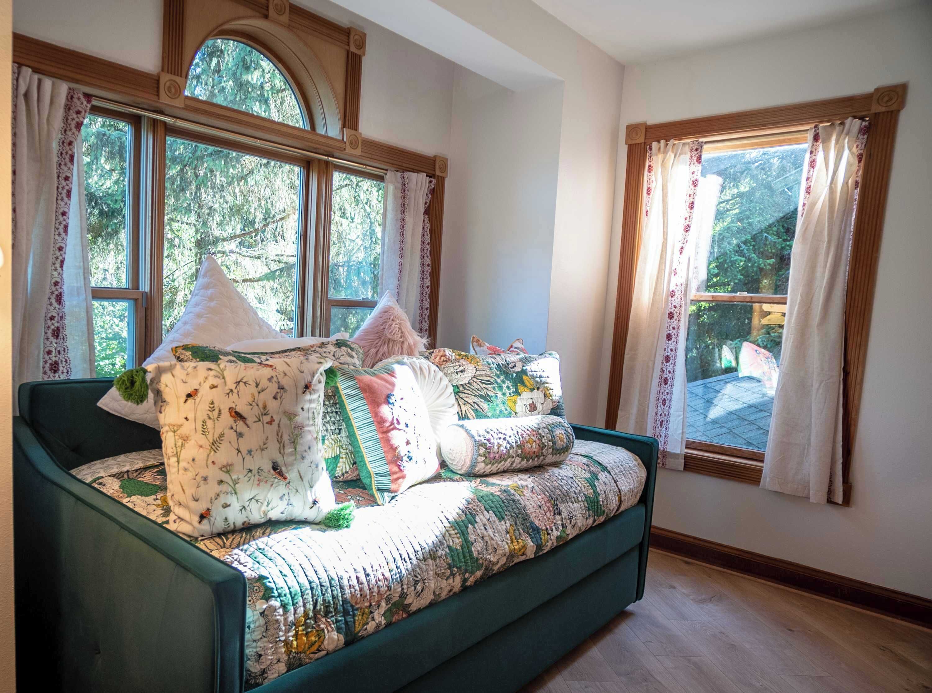 A cozy room with a green sofa bed adorned with floral and patterned blankets, large windows with curtains, and a view of greenery outside.