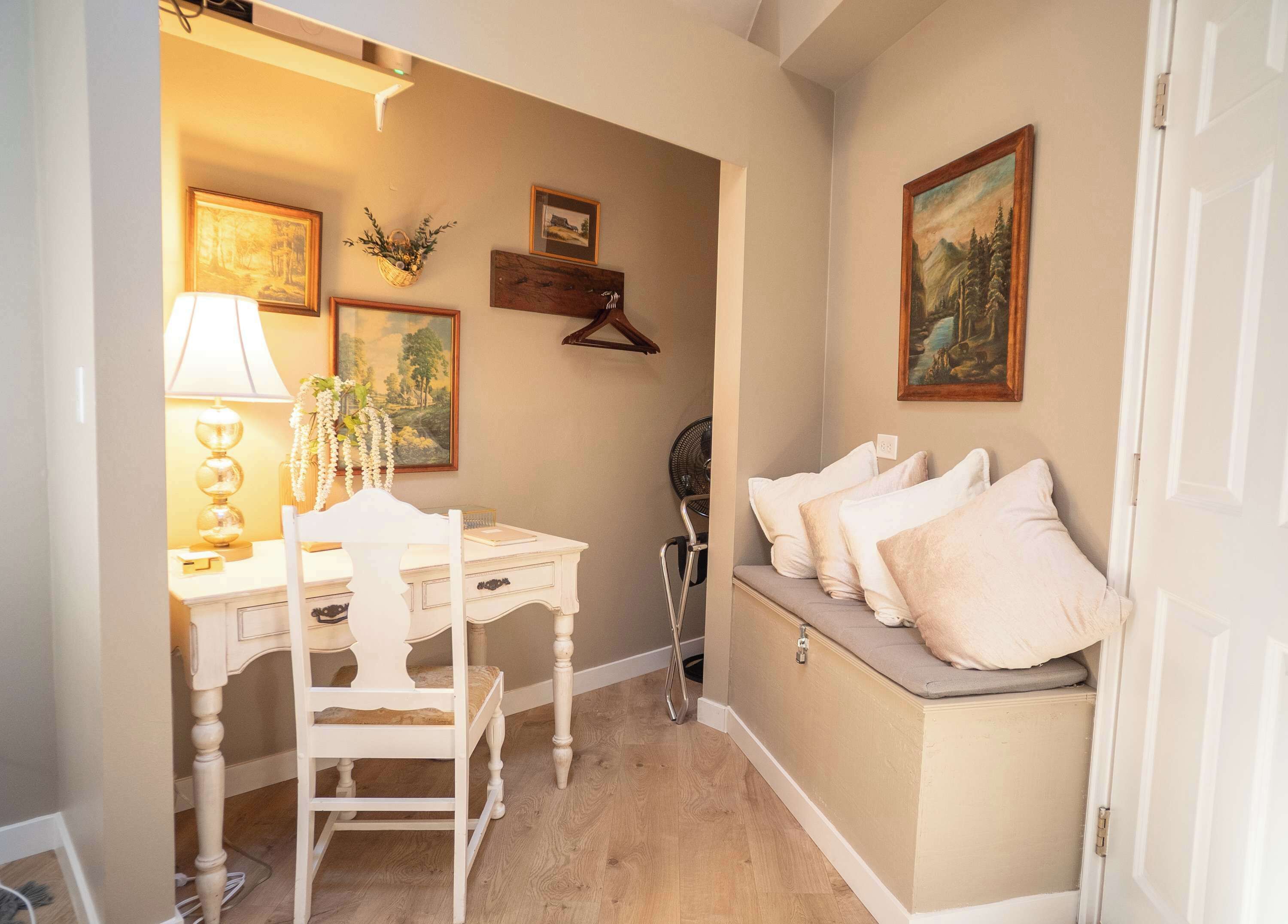 Une pièce chaleureuse avec un bureau blanc, une chaise et un banc agrémenté de coussins. Les murs sont ornés de tableaux encadrés, et un panier rempli de plantes vertes est posé dans un coin.