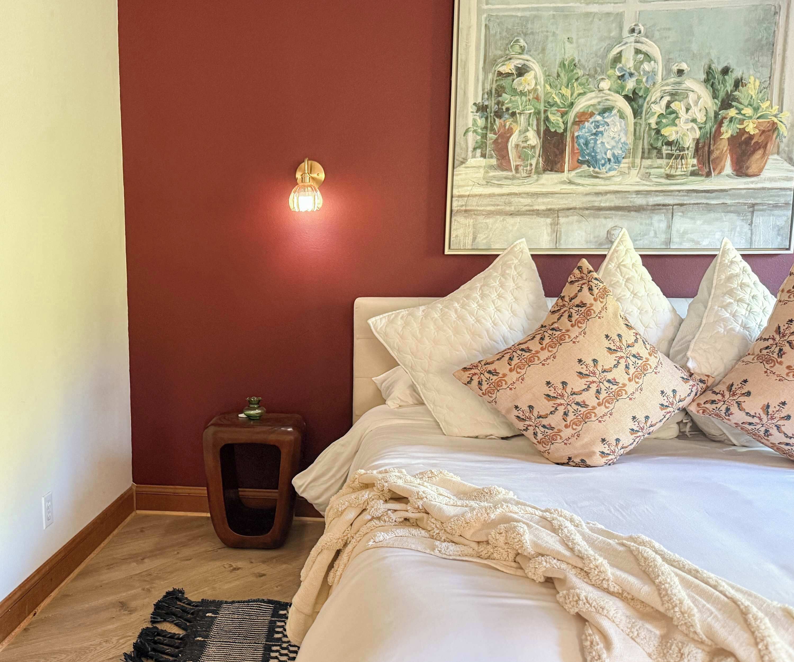 Un dormitorio con cama, almohadas, una mesita de noche, una lámpara y un cuadro en la pared.