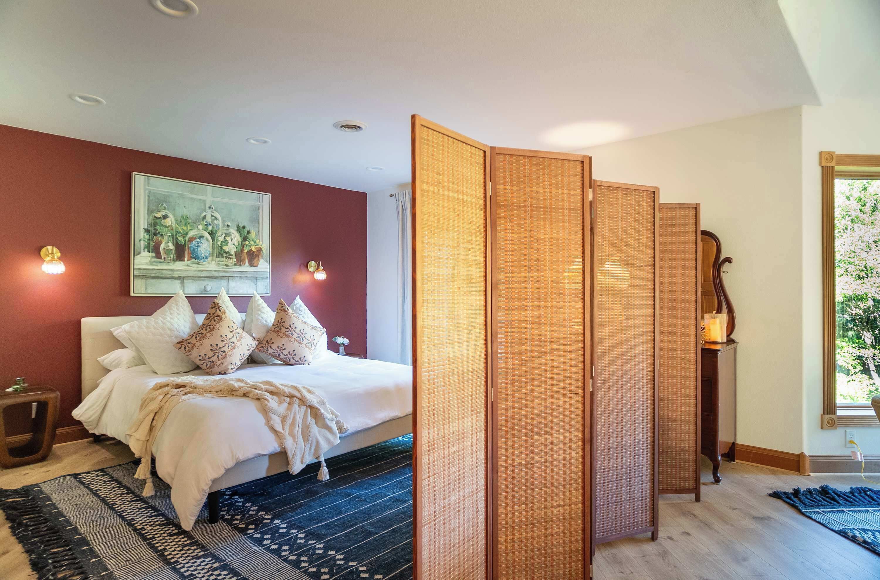 A bedroom with a bed, a painting, a wooden screen, and a window with a view.