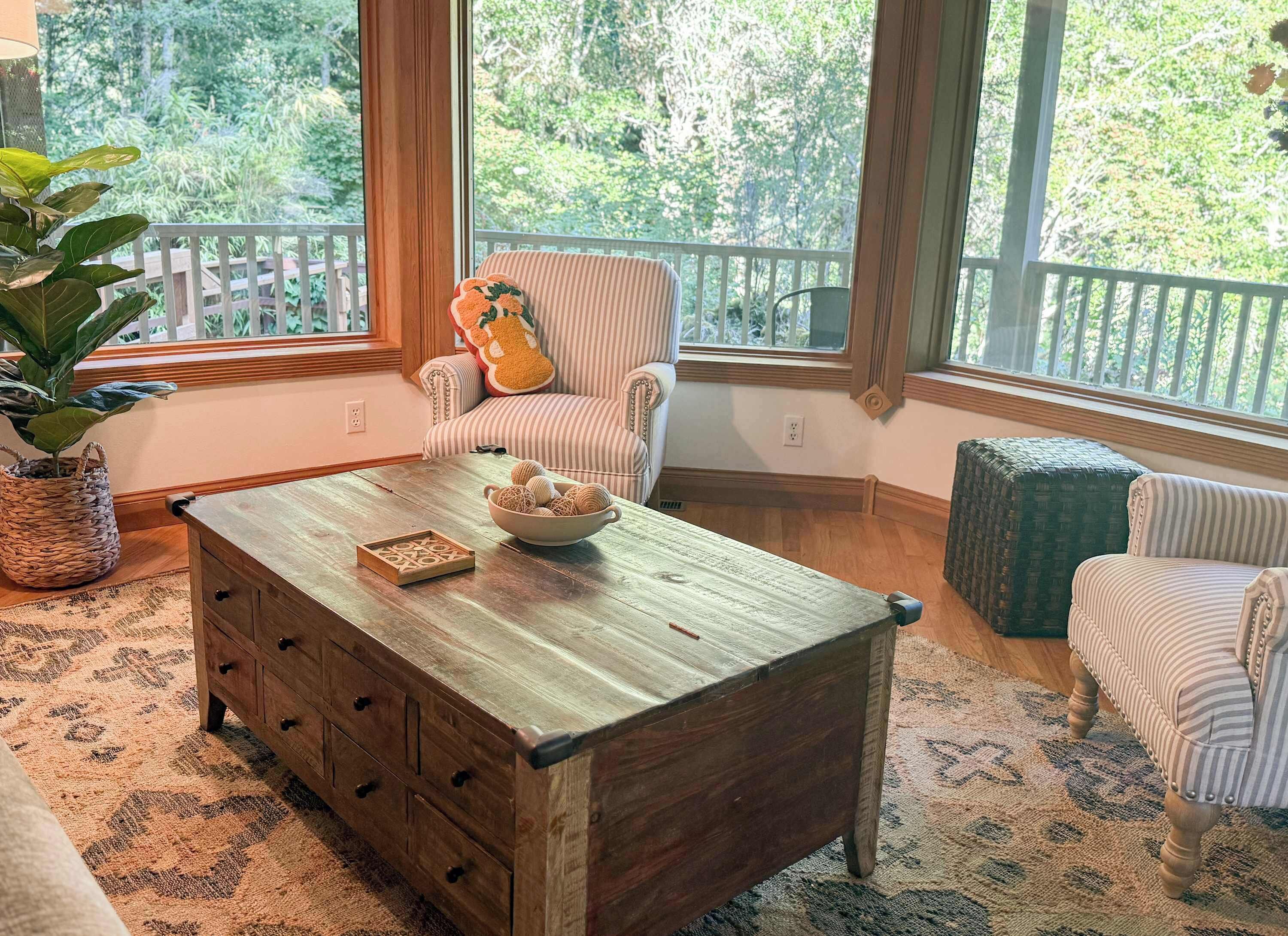 Quarto aconchegante com mesa de centro de madeira, poltronas listradas e uma janela grande com vista para árvores.
