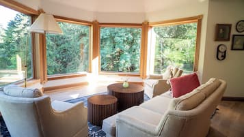 Una sala de estar acogedora con un sofá color beige, dos mesitas de madera redondas y un ventanal con vistas a árboles.