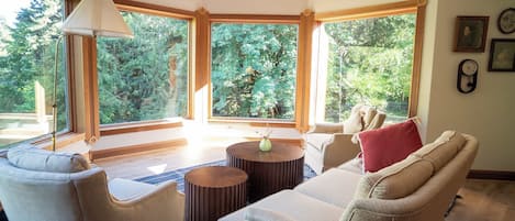 A cozy living room with a beige sofa, two round wooden side tables, and a large window offering a view of trees.