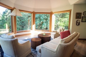 Una sala de estar acogedora con un sofá color beige, dos mesitas de madera redondas y un ventanal con vistas a árboles.