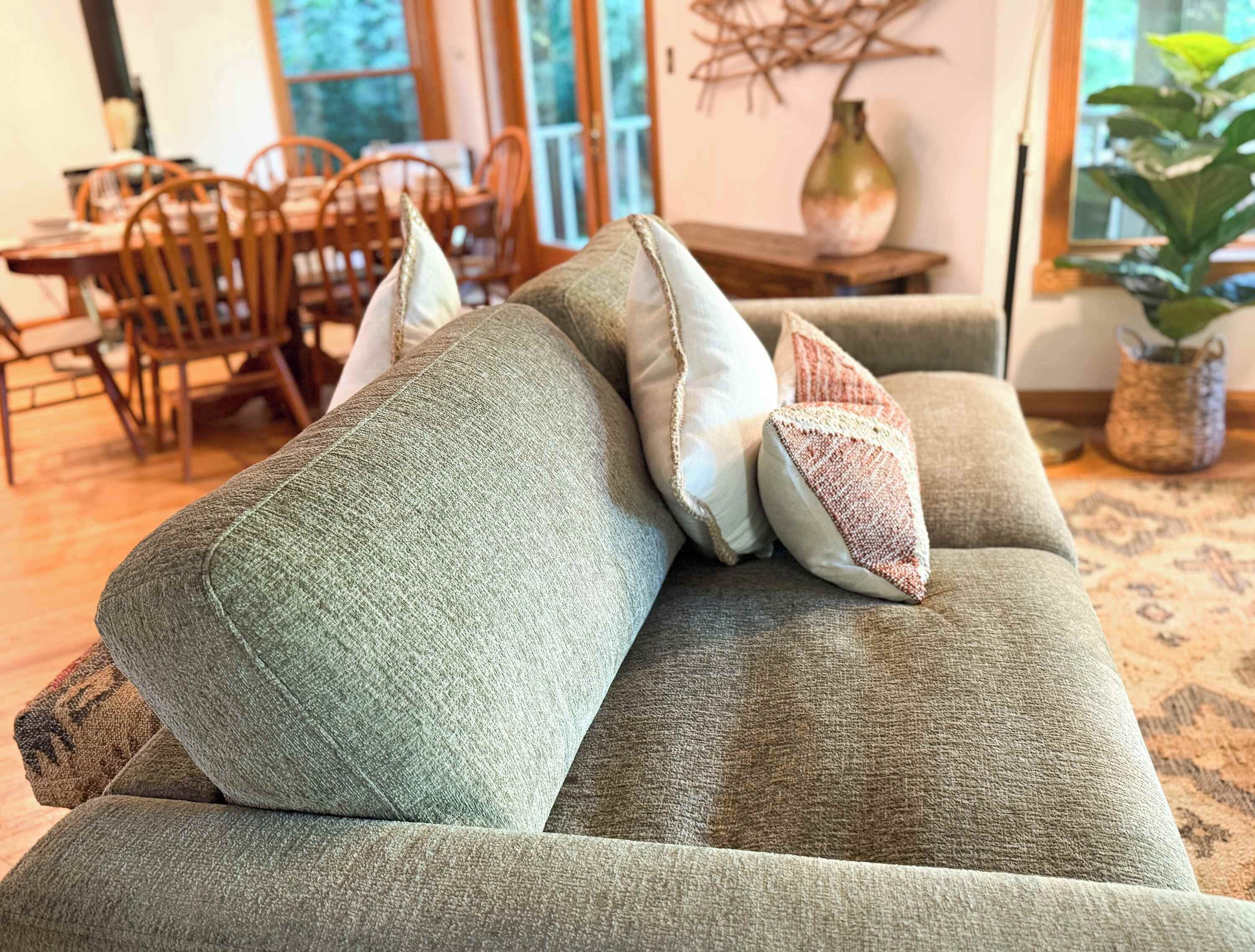 A green sofa with multiple cushions in a well-lit room with a dining area in the background.