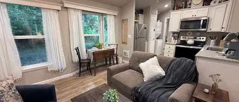 A living room with a sofa, coffee table, and a dining area with a table and chairs. There is a kitchen with white cabinets and appliances.