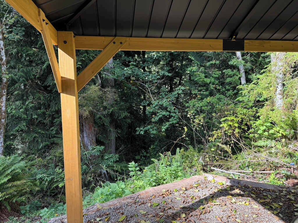 Une structure de pergola en bois, dotée d’un toit et entourée d’une végétation forestière dense.
