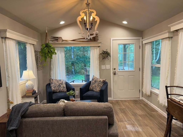 A cozy living room with a sofa, two armchairs, a chandelier, and large windows offering a view of the outdoors.