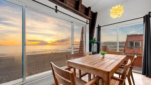 A spacious room with a large wooden table and chairs, a potted plant, and a view of the beach at sunset through large windows.
