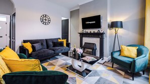 Un salón moderno con un sofá negro, un sillón azul turquesa y una mesa de centro con una revista. Un televisor de pantalla plana está montado sobre una chimenea.