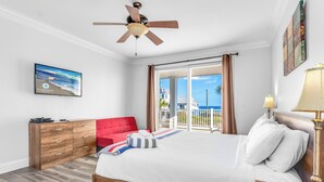 Ein Schlafzimmer mit Bett, Deckenventilator, Holzkommode, rotem Sessel, Fenster mit Vorhängen und Meerblick.