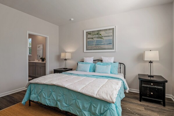 A bedroom with a bed, two bedside tables, a nightstand, a framed picture on the wall, and a bathroom visible through an open door. - NEW Coastal Retreat • Fire Pit • Golf Escape • 3 mi to Holden Beach • Sleeps 6 (Supply)