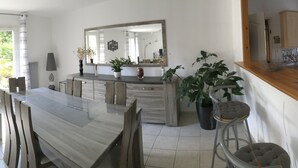 A modern kitchen with a large island, a dining area with a glass table, and a view of the outdoors. - Contemporary, single-storey house (Corme-Royal)