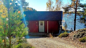 Une petite cabane à un seul étage, avec un toit sombre, une façade rouge et une porte blanche. Elle est située sur un chemin de gravier, avec des arbres et une étendue d’eau en arrière-plan.