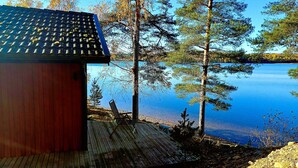 Une terrasse en bois mène à un chalet doté d’un toit en tuiles, qui offre une vue sur un lac paisible entouré d’arbres.