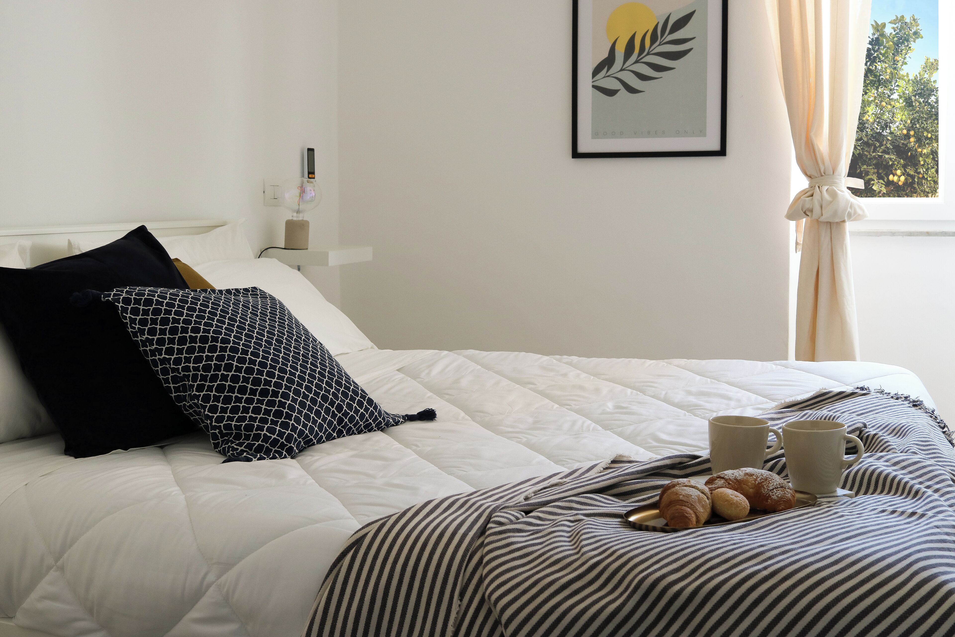 A neatly made bed with a white duvet, black and white striped blanket, and two black pillows. There are two white mugs and a plate with croissants on the bed.