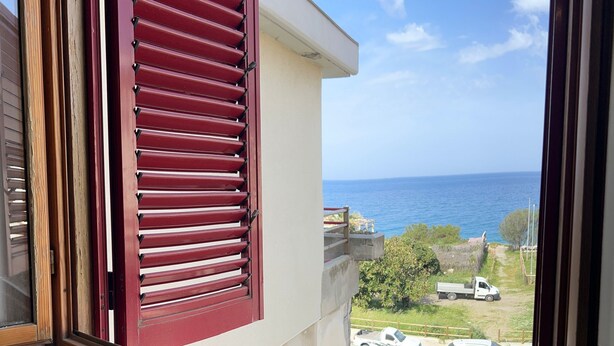 A window with red shutters overlooking a street with parked cars and a clear view of the sea. - Sailor's House (Milazzo)
