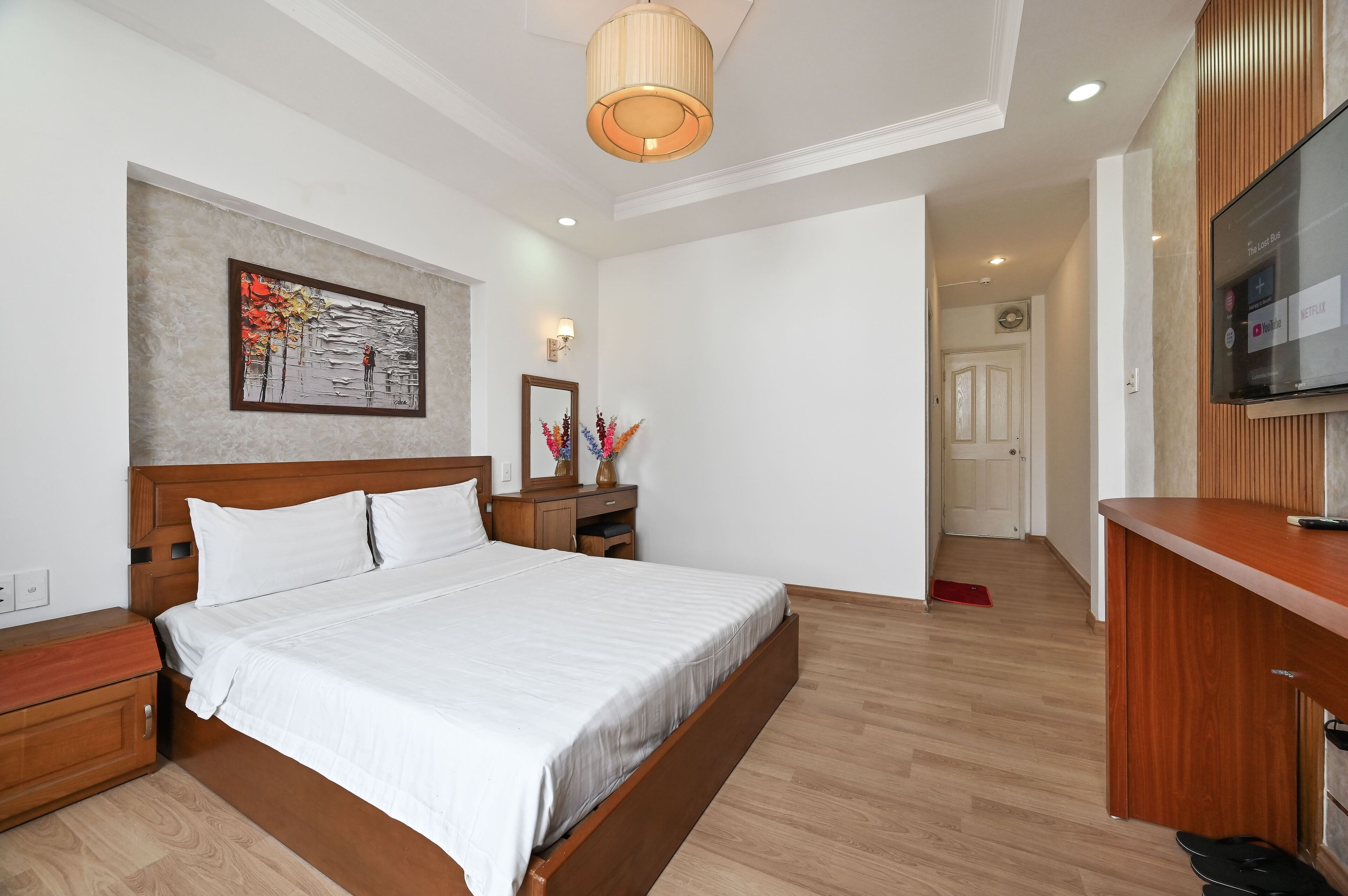 Una habitación de hotel moderna con una cama grande, cabecero de madera, un cuadro en la pared, una mesita de noche con una lámpara y un televisor montado en la pared.