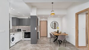 A modern kitchen with grey cabinets, a microwave, and a washing machine. There is a dining area with a table and chairs, and a wooden door in the background. - Acropolis Oasis - 2BR, Koukaki (Athina)