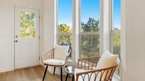 A room with a door, two wicker chairs with cushions, a small table, and large windows offering a view of trees and a clear sky.