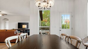 A dining area with a large wooden table and chairs, a chandelier, and a view of the outdoors through a door.