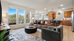 A spacious living area with a brown leather sofa, a grey armchair, and a round coffee table. There are large windows offering a view of greenery outside.