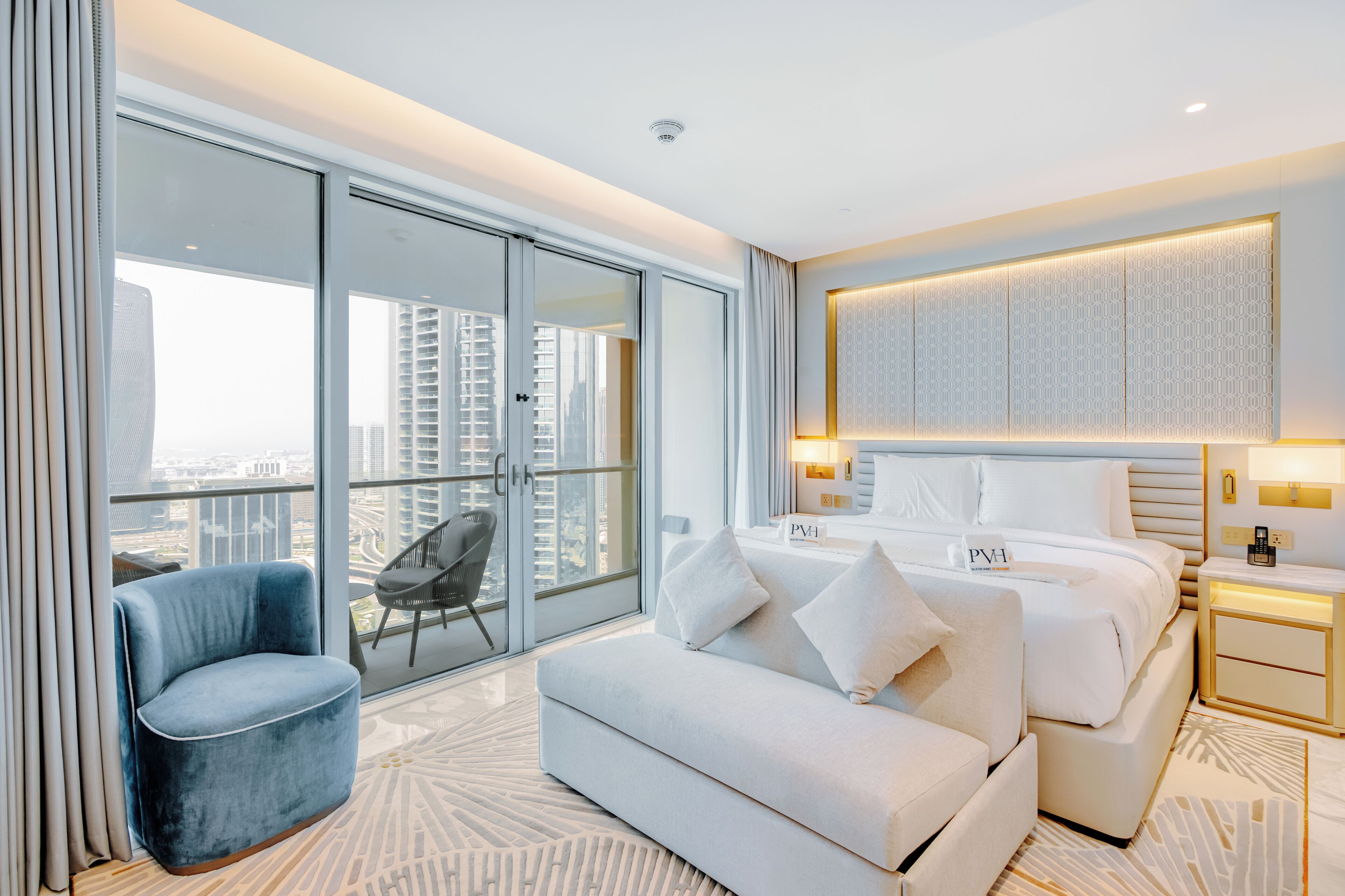 Une chambre d’hôtel moderne dotée d’une grande porte-fenêtre coulissante donnant sur un balcon, d’un lit avec une literie blanche, d’un fauteuil bleu et d’une petite table de chevet avec une lampe.