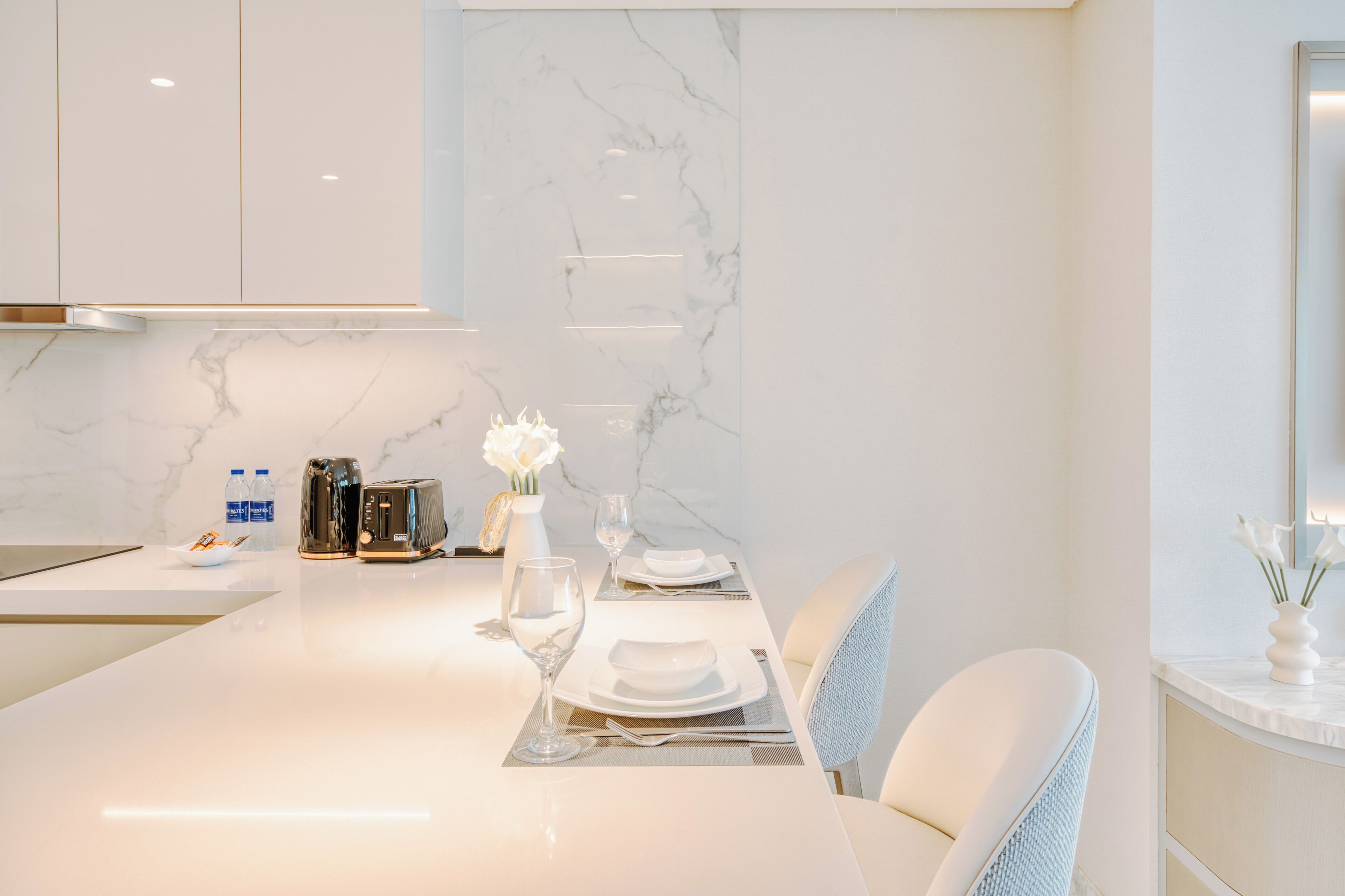 Une cuisine moderne avec un pare-feu en marbre, des meubles blancs et un coin repas avec une table blanche mise pour deux personnes.