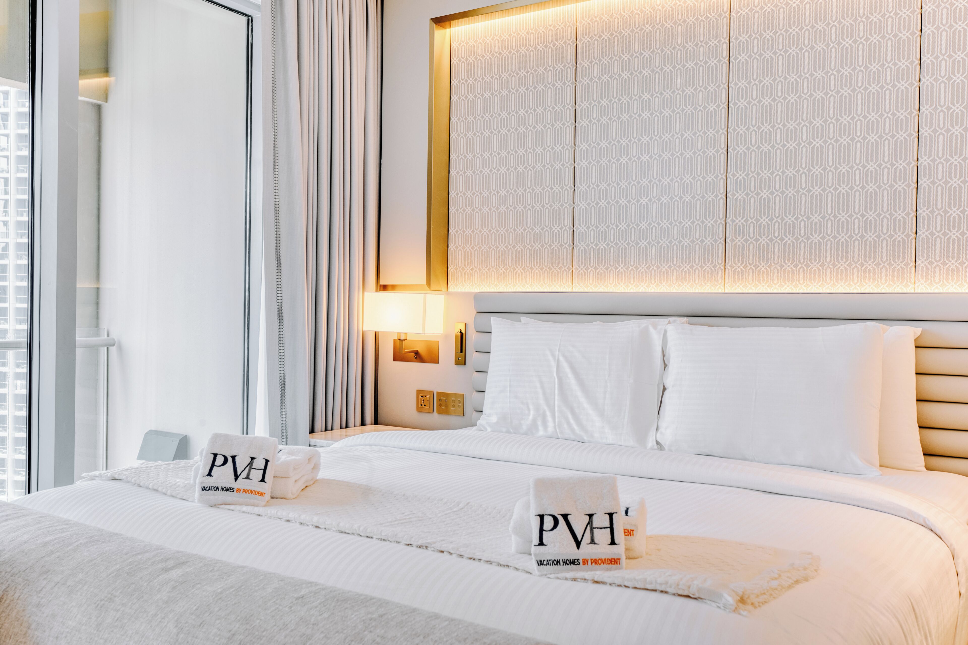 Une chambre d’hôtel avec un lit bien fait, une grande fenêtre avec des rideaux, une lampe fixée au mur et une tête de lit à motifs.
