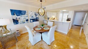 A dining area with a round table, white chairs, and a chandelier. There are abstract paintings on the wall and a kitchen in the background. - Stunning Gulf Front  Mainsail 143 with Free Beach Service for 2 (Miramar Beach)