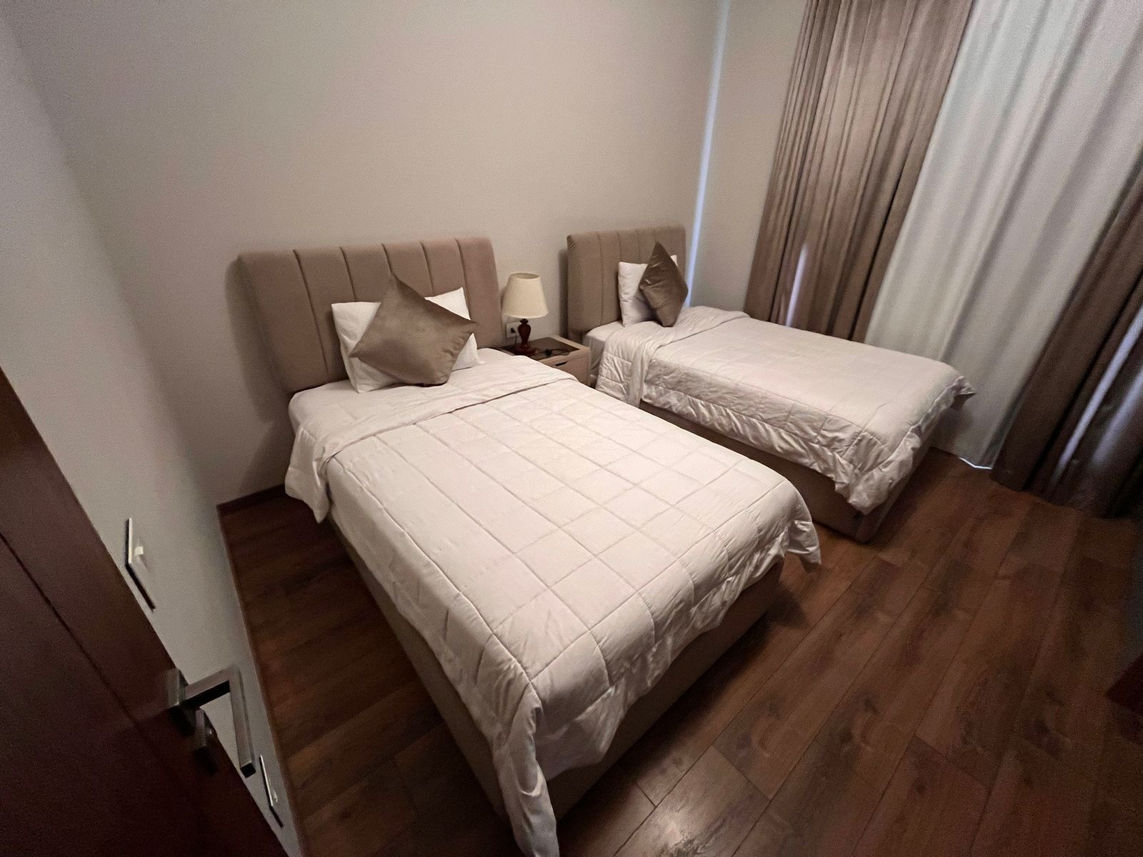 A hotel room with two beds, a wooden floor, and a window with curtains.