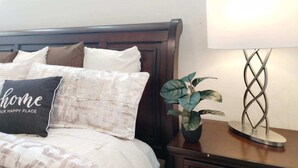 Una cama bien hecha con un cojín que dice 'hogar, tu lugar feliz', una planta en maceta sobre una mesita de noche y una lámpara de diseño peculiar.
