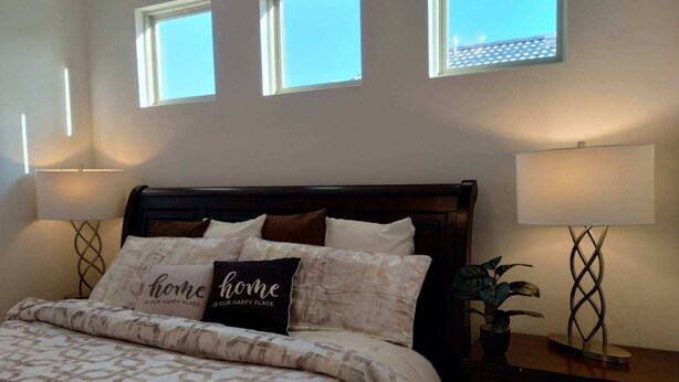 Un dormitorio con una cama, dos lámparas de noche, una mesita de noche con una planta y tres ventanas.