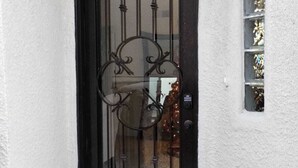 Una reja de metal negro con detalles decorativos, una pared blanca con textura y un ventanal con estantes.