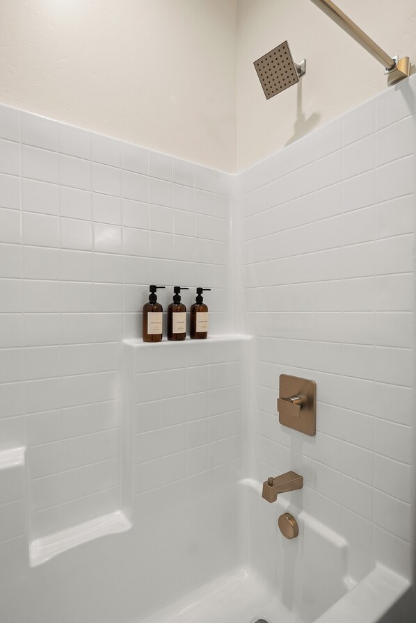 A bathroom with white tiled walls, a showerhead, and three bottles of toiletries on a shelf.