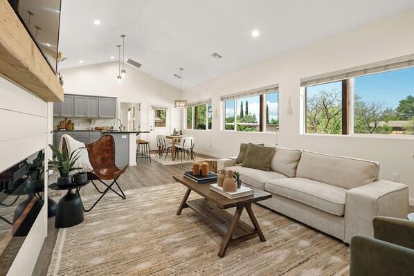 A spacious living area with a sofa, coffee table, and a dining table. There are large windows providing natural light and a view of the outdoors.