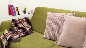 A green sofa with patterned pillows, a framed artwork on the wall, and a shelf with books and a cactus.