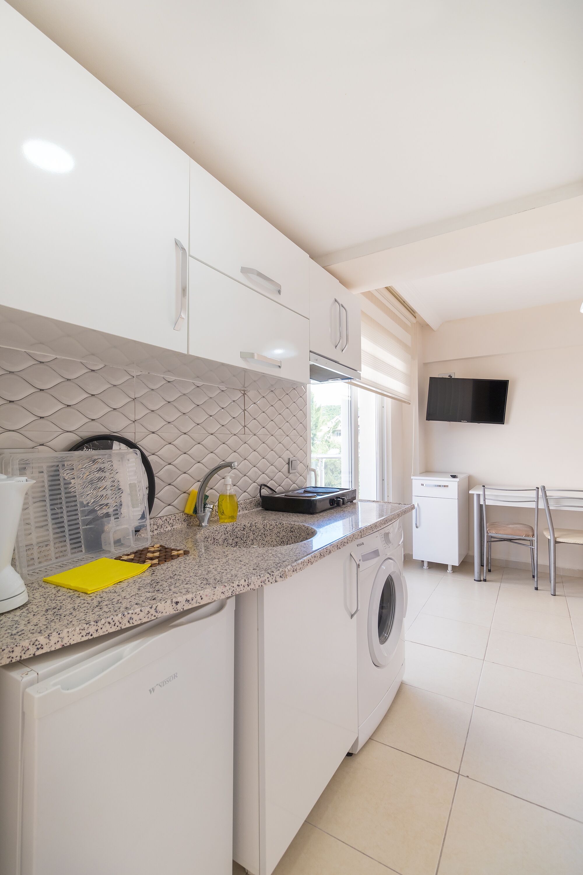 A kitchen with a washing machine, a small refrigerator, a microwave, and a TV mounted on the wall.