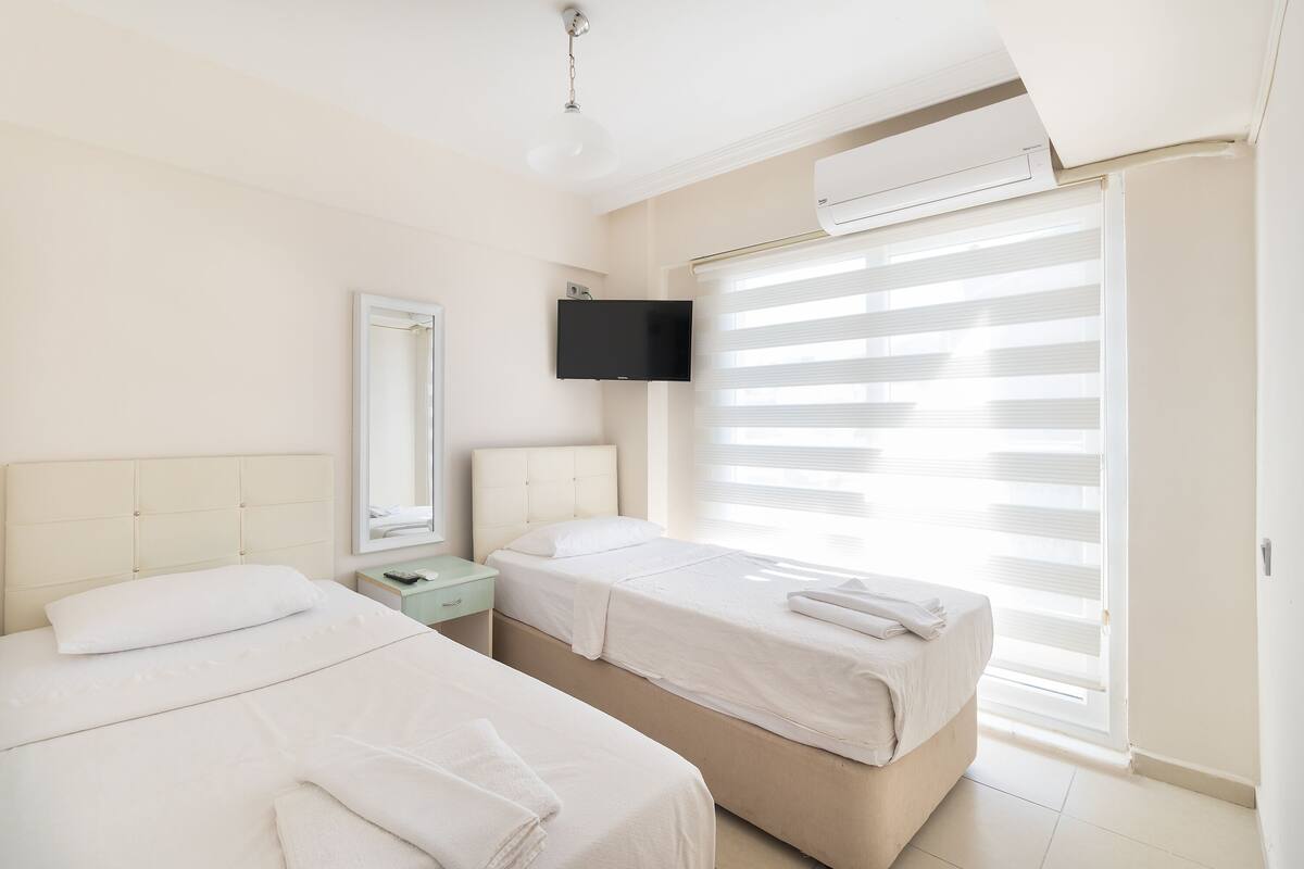 A hotel room with two beds, a flat-screen TV, a large window with blinds, and an air conditioning unit.
