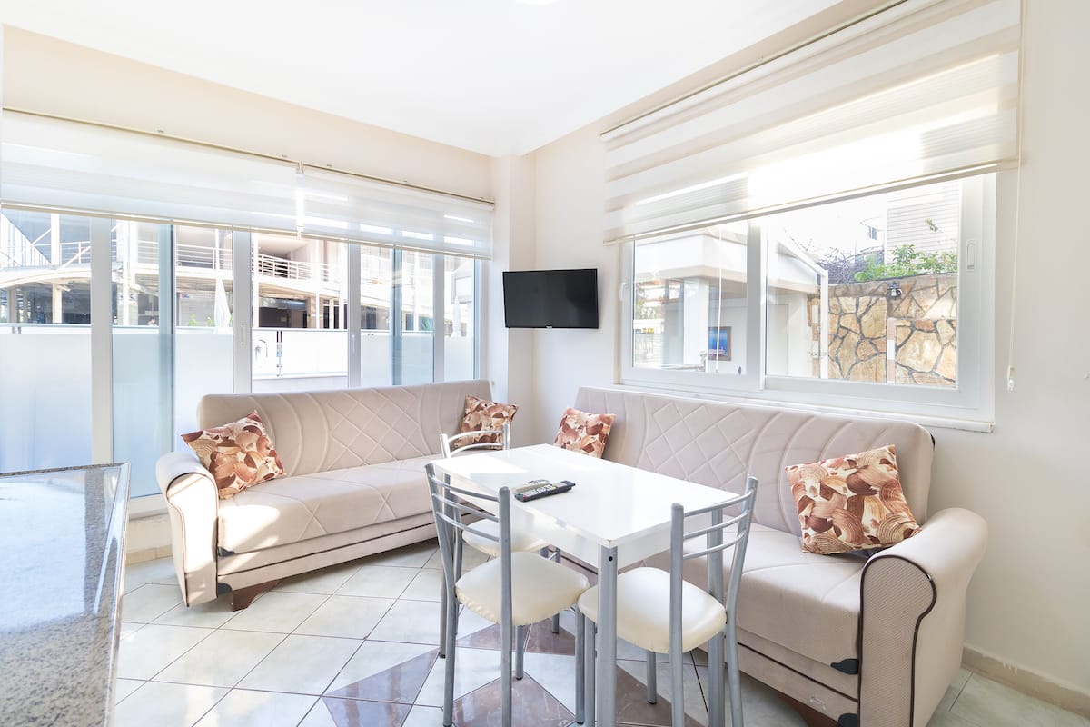 A modern living room with a sofa, a dining table, and chairs. There is a flat-screen TV mounted on the wall.