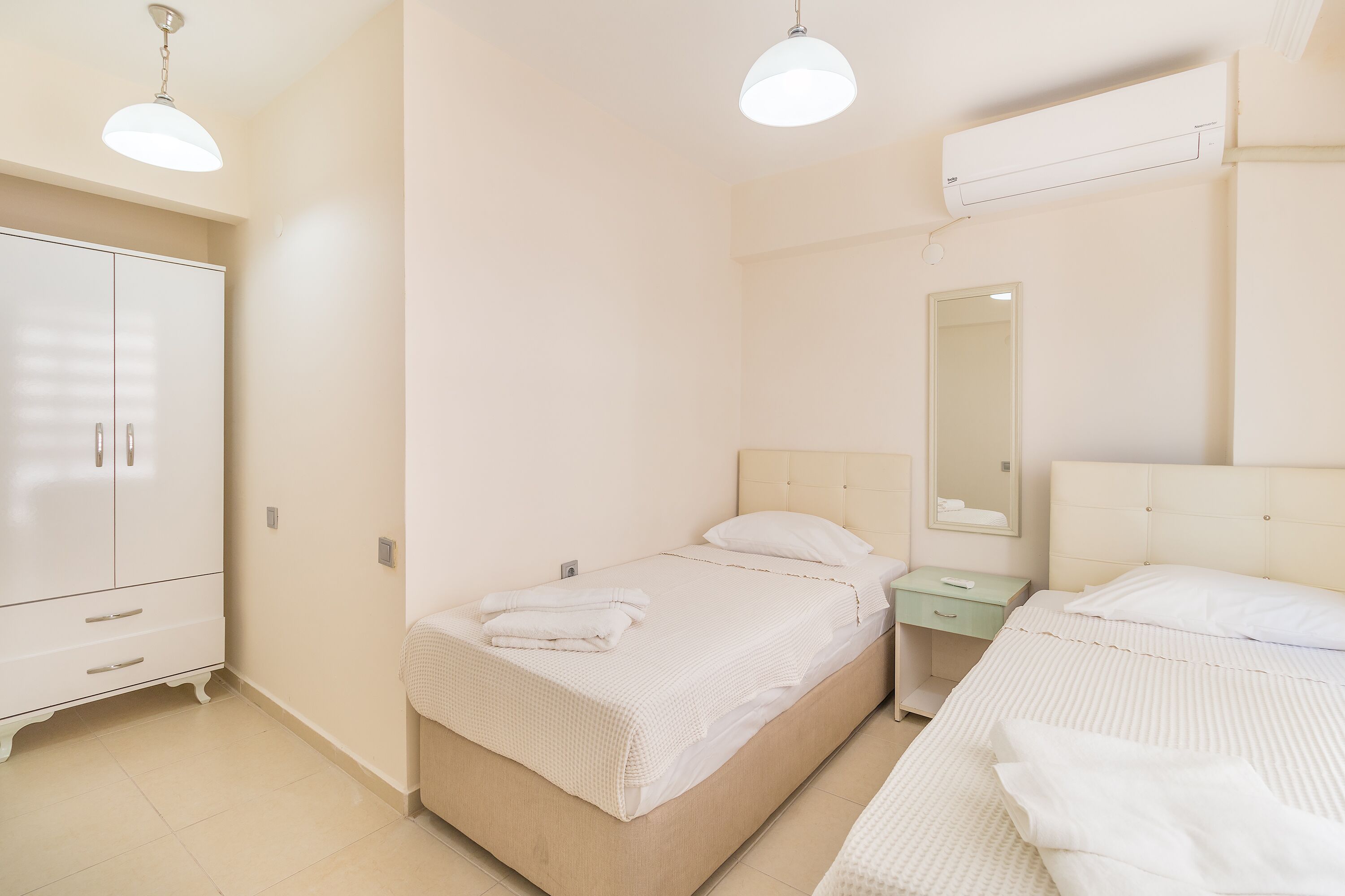 A bedroom with two beds, a wardrobe, a bedside table, a mirror, and an air conditioning unit.